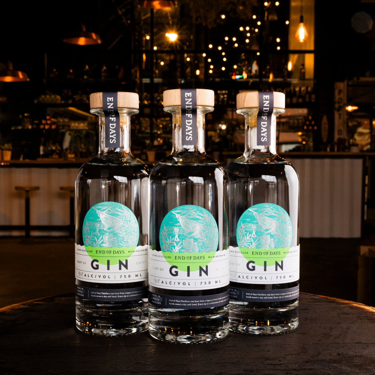 Three bottles of Port of Entry Gin lined up on a bar table with warm lights behind